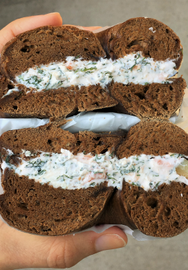 Hand holding two stacked halves of a pumpernickel bagel with salmon dill cream cheese