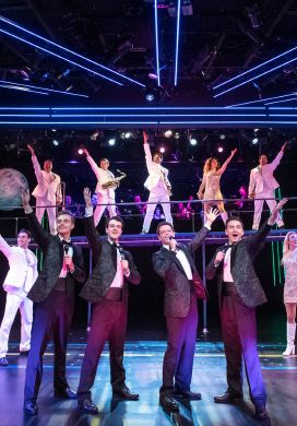 Four men in black suits singing with a backup ensemble of actors dressed in white