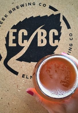 Top down view of hand holding beer over a table with the logo for Elm Creek Brewing Co.
