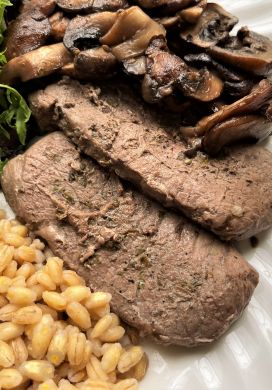 Close up of venison steak on a white plate with barley, mushrooms, and mixed greens