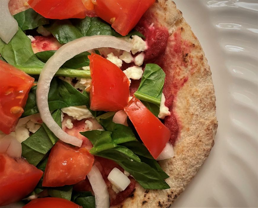 Pink Beetroot Pesto Sauce pita pizza Close up of a pita topped with pink beet pesto, spinach, onions, fresh tomatoes, and crumbled feta