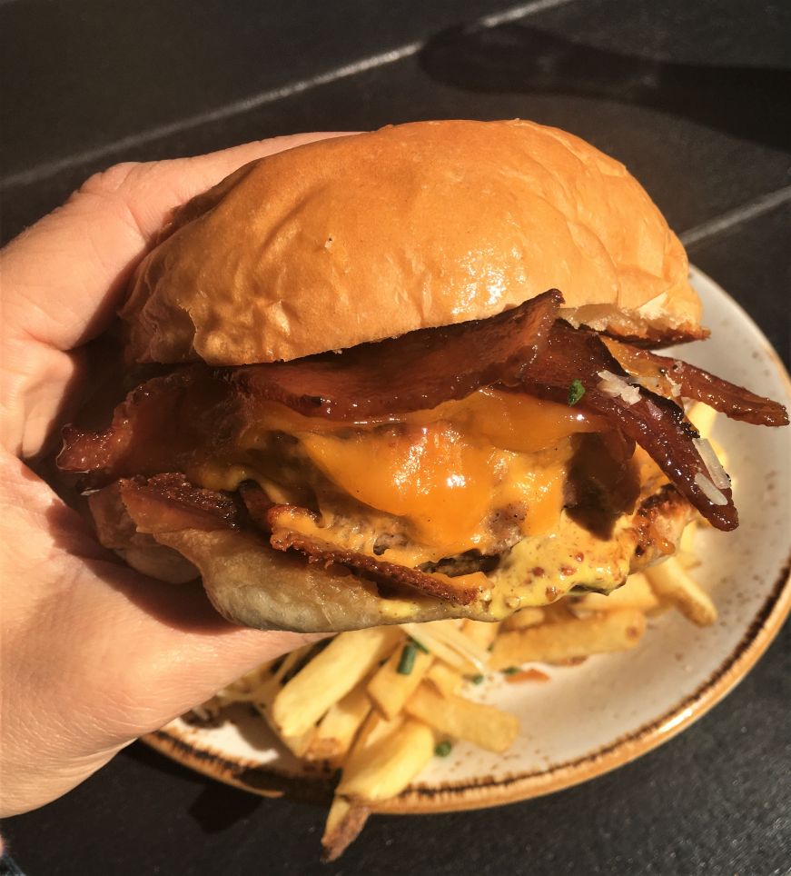 Hand holding bacon cheeseburger with a plate of fries in the background