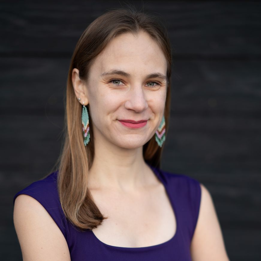 Headshot of Stacy, a white woman with brunette hair wearing a purple shirt and beaded earrings