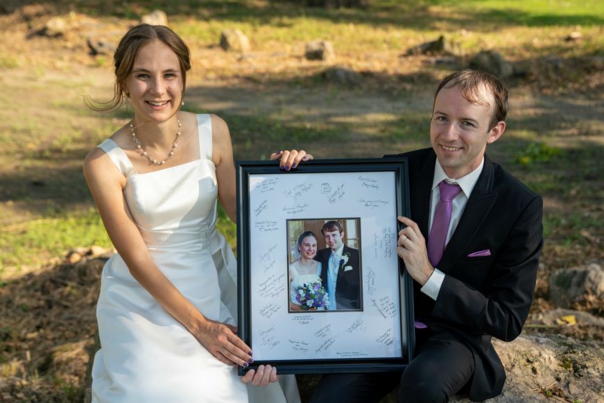 Stacy in a wedding dress and Mike in a tux holding their framed wedding photo Stacy in a wedding dress and Mike in a tux holding their framed wedding photo