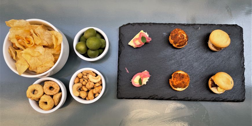 Top down view of a plate of miniature sandwiches and small bowls filled with chips, olives, and nuts