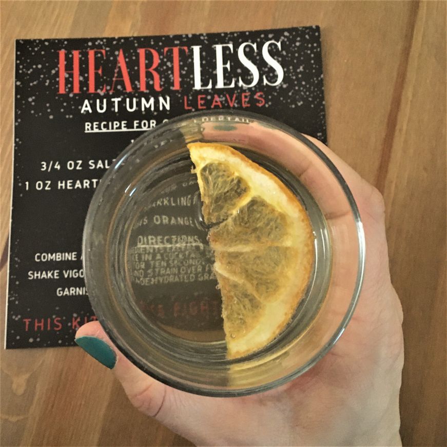 Autumn Leaves cocktail, Heartless Top down view of a hand holding a light orange cocktail garnished with a dried grapefruit slice