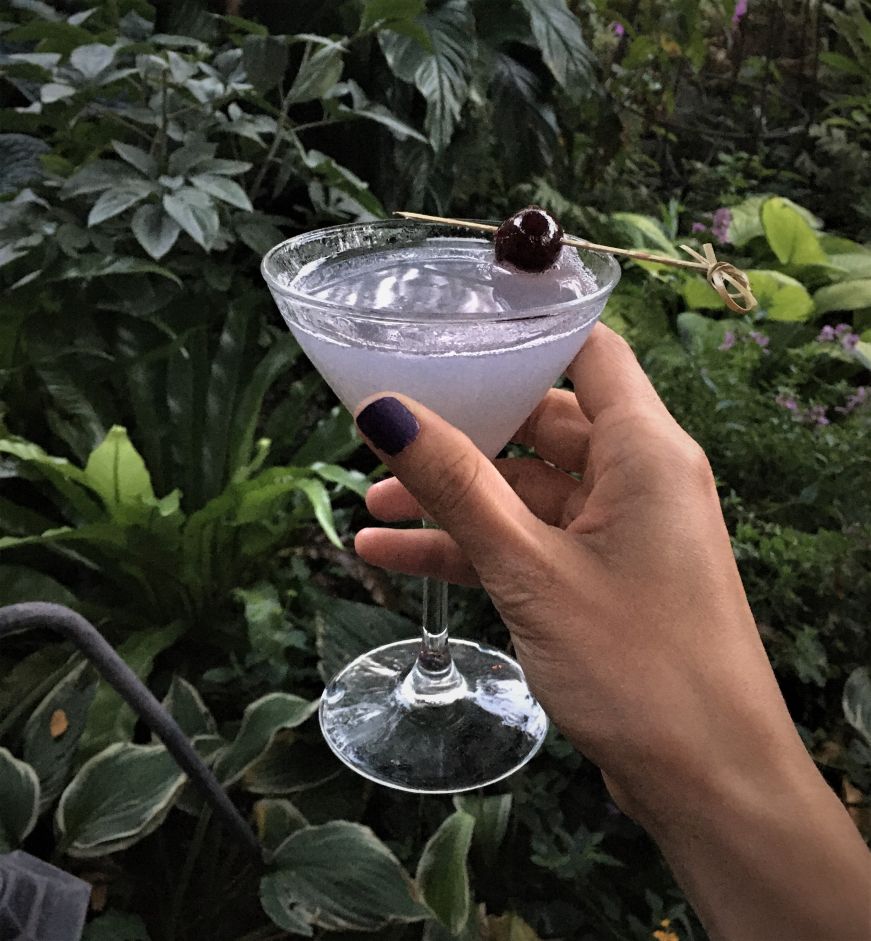 Hand holding a purple cocktail garnished with a cherry