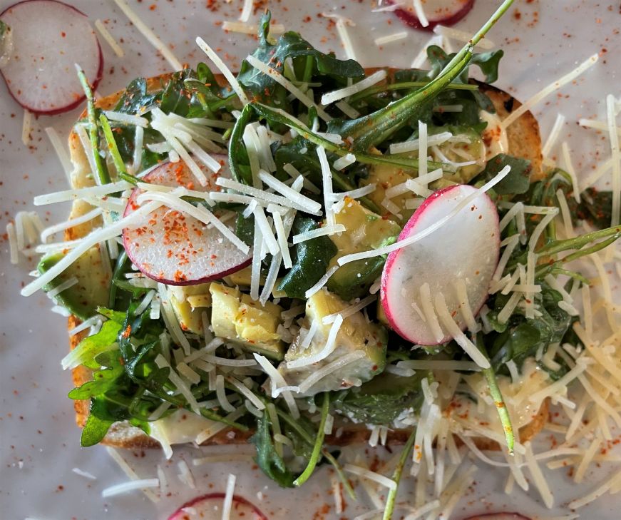 Piece of toast loaded with arugula, sliced radishes, chunks of avocado, and shredded cheese