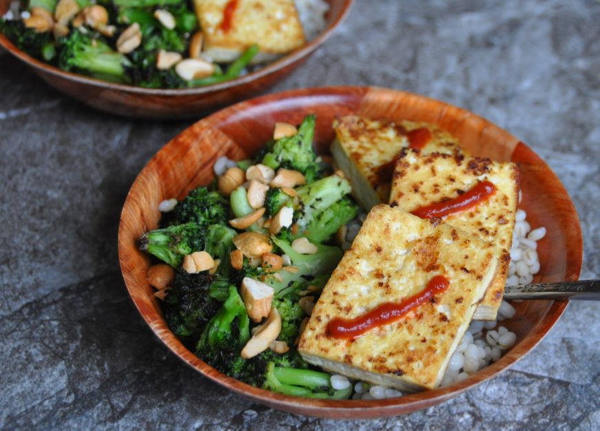 Two wooden bowls filled with barley topped with roasted broccoli, tofu, sriracha sauce, and chopped cashews