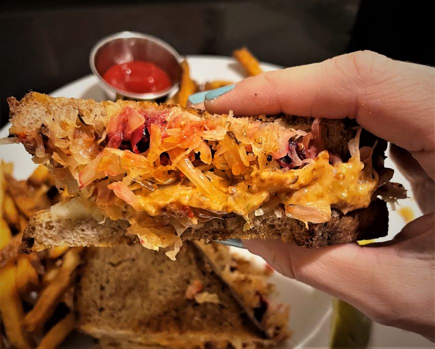Hand holding a toasted sandwich filled with beets, sauerkraut, and melted cheese with a plate of fries in the background