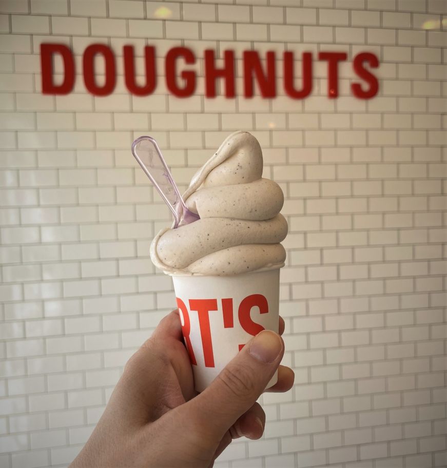 Stacy's hand holding a cup of soft serve with a sign reading "Doughnuts" in the background