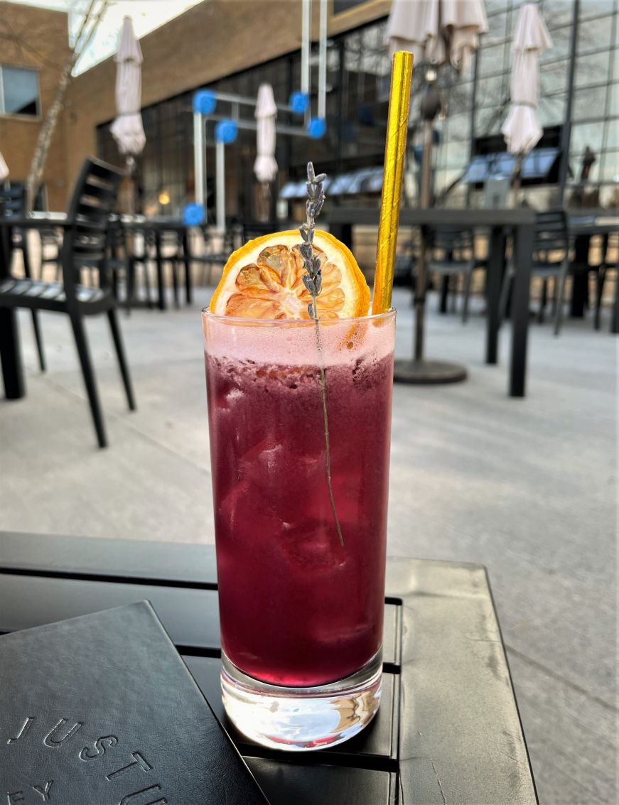 Purple cocktail garnished with a sprig of lavender and a dried citrus slice sitting on an outdoor table