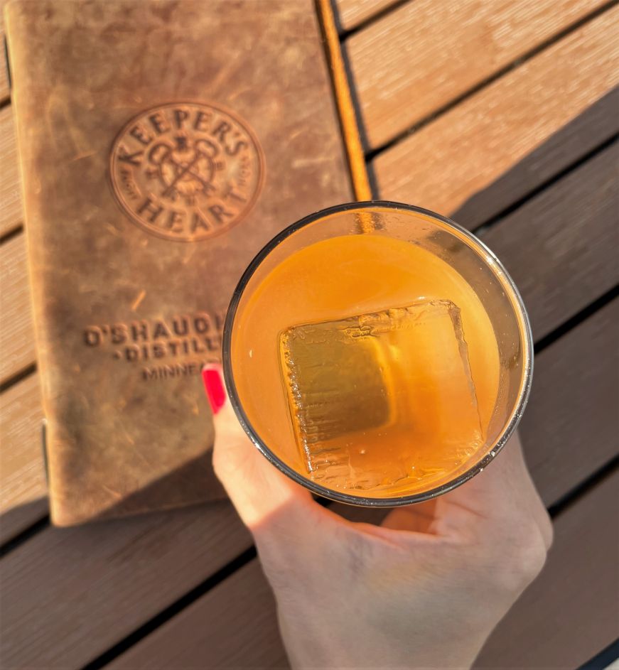 Stacy's hand holding an amber-colored cocktail in a rocks glass with a large square ice cube