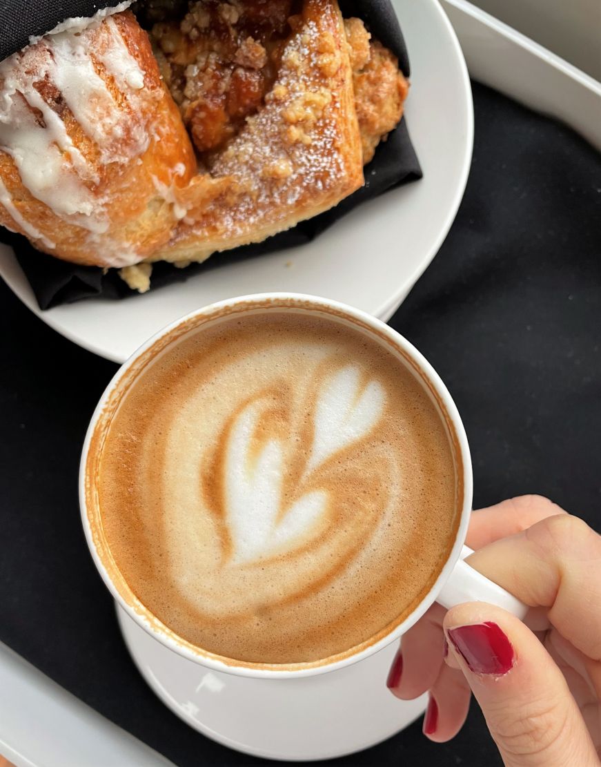 Hand holding a cappuccino with latte art with a basket of pastries in the background
