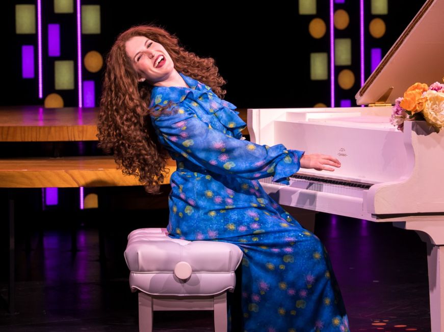 Woman in a blue dress with long curly hair playing a piano and singing