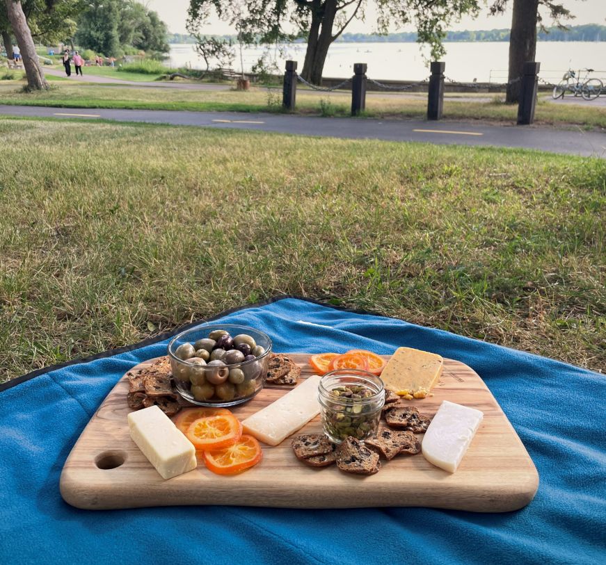 Cheese board on a blue picnic blanket with a path and lake in the background