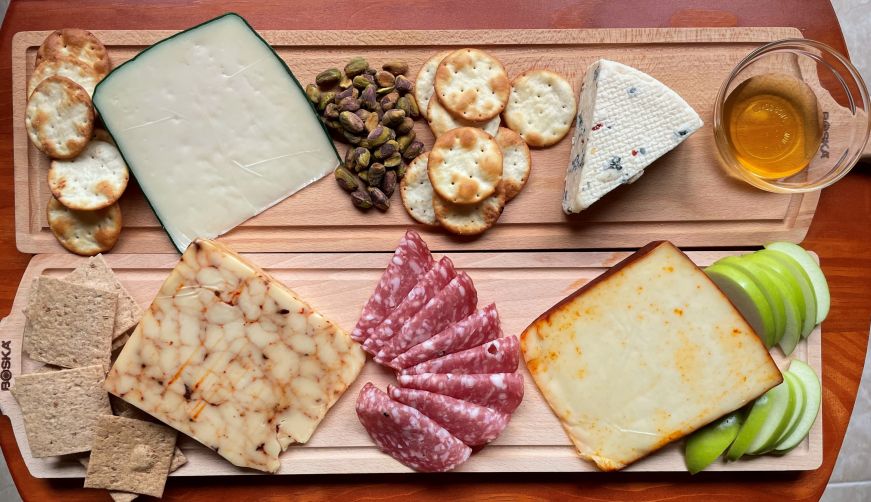 Two rectangular cheese boards with four blocks of cheese, sliced apples, salami, pistaschios, crackers, and a small bowl of honey