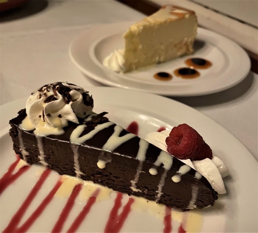 Slice of chocolate torte on a white plate drizzled with raspberry sauce with a slice of vanilla cheesecake in the background