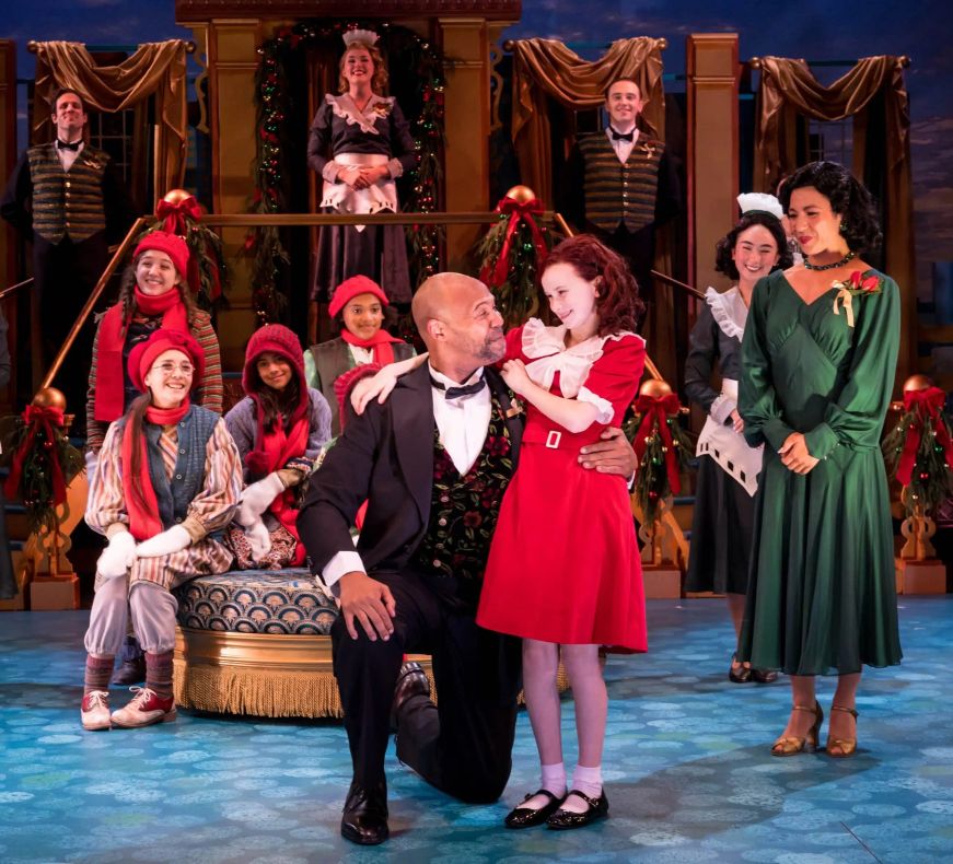 Red headed girl being embraced by a man in a tuxedo, surrounded the rest of cast in Christmas-themed costumes