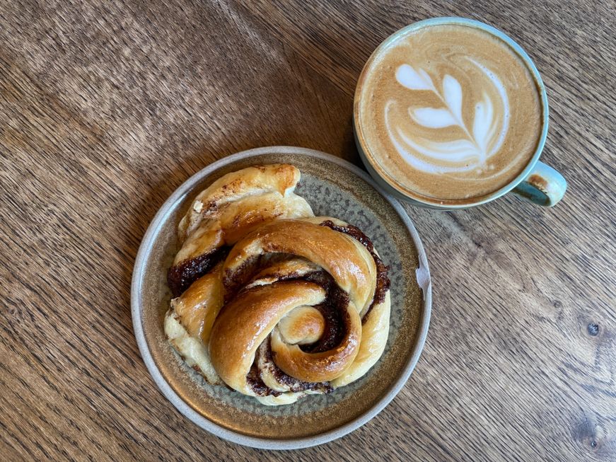 Cinnamon roll, Hygge Coffee & Micro Bakery Top down view of a twisted cinnamon roll and a cappucino with latte art
