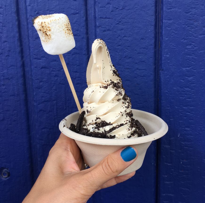 Hand holding cardboard bowl filled with coffee soft serve topped with chocolate dust and garnished with a toasted marshmallow and Oreo cookies