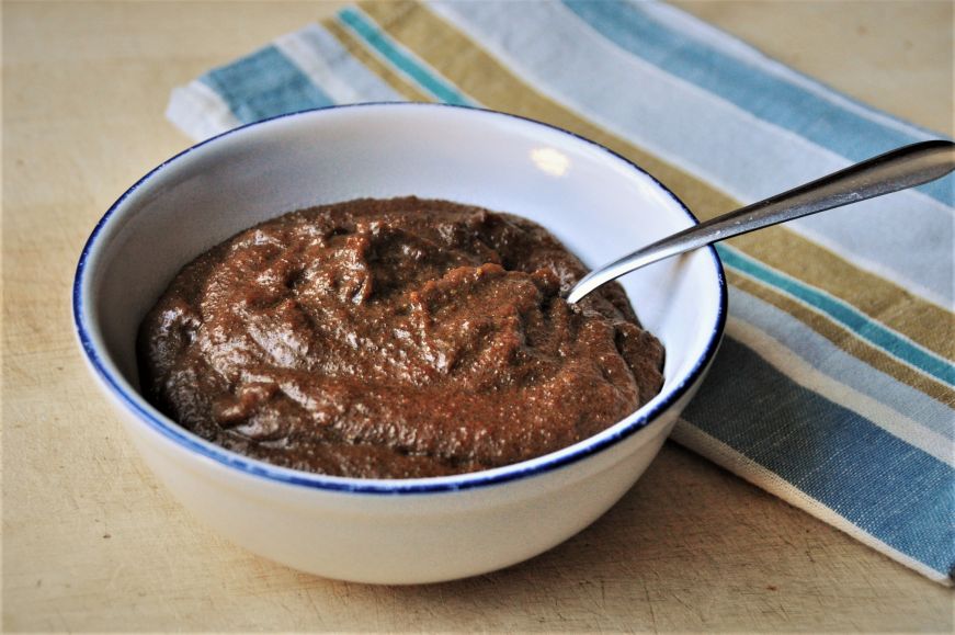 Bowl of cornmeal molasses pudding with a spoon in it and a striped napkin the background