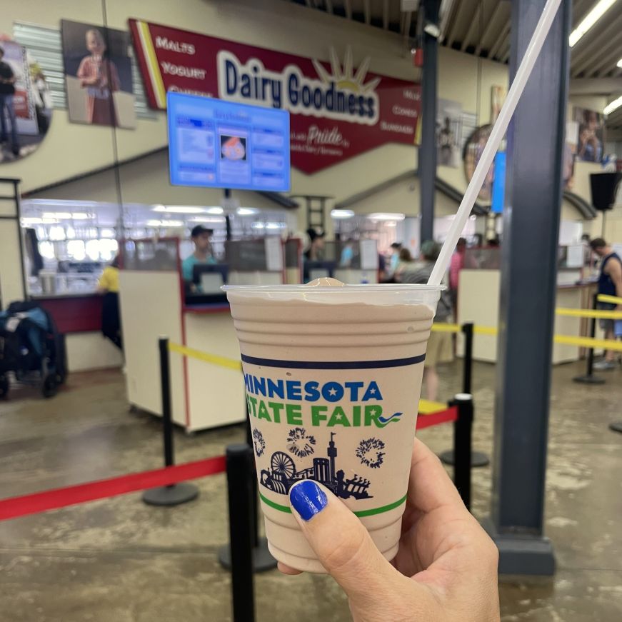 Hand with blue nail polish holding a chocolate malt with the Dairy Goodness booth in the background