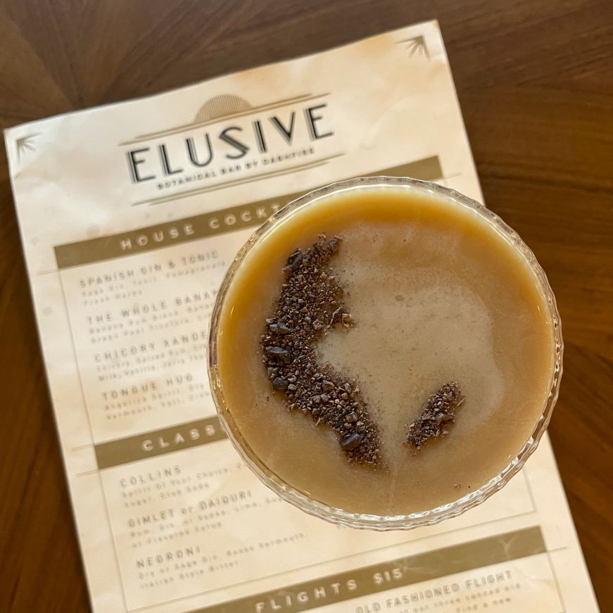 Top down view of a brown, creamy cocktail garnished cocoa nibs with a cocktail menu in the background