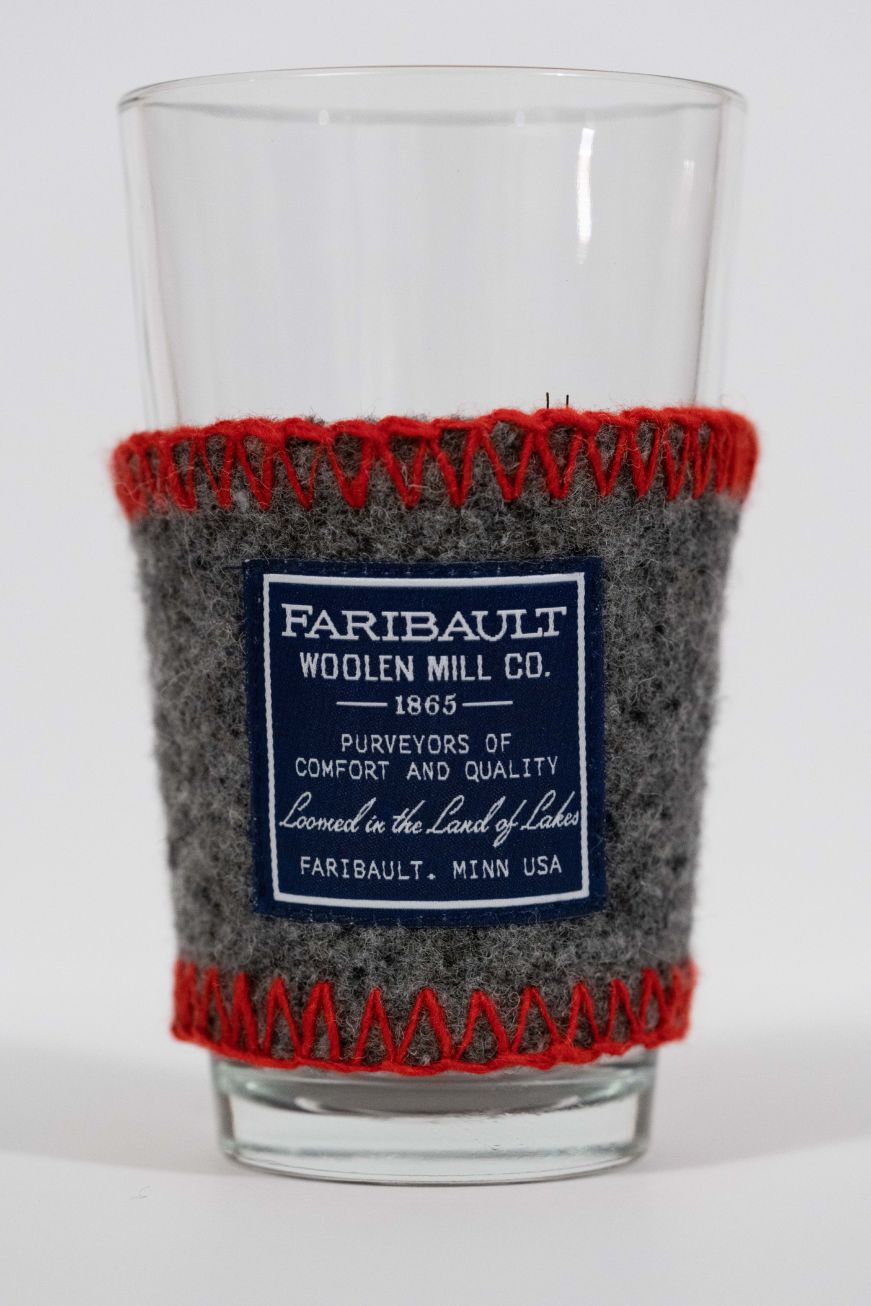 Pint glass wrapped with a gray woolen sleeve with red stitching and a Faribault Woolen Mill label