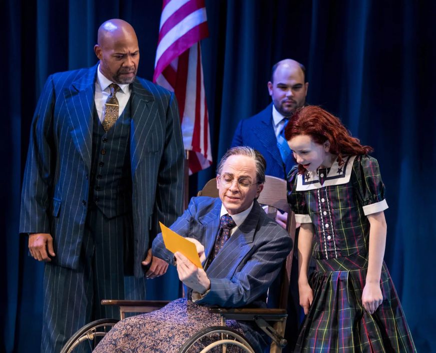 Man in a wheelchair showing a document in a red-headed girl, surrounded by men in suits