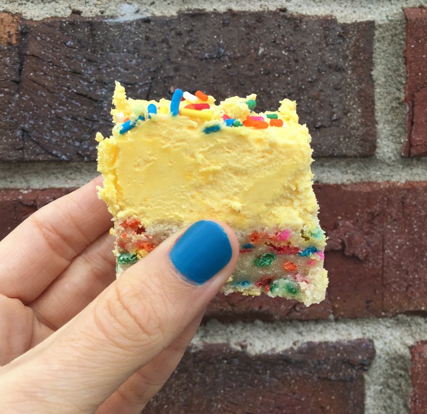 Side view of a hand holding a funfetti cookie bar with a thick layer of yellow frosting