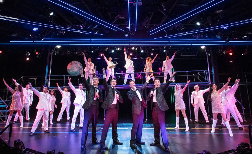 Four men in black suits singing with a backup ensemble of actors dressed in white