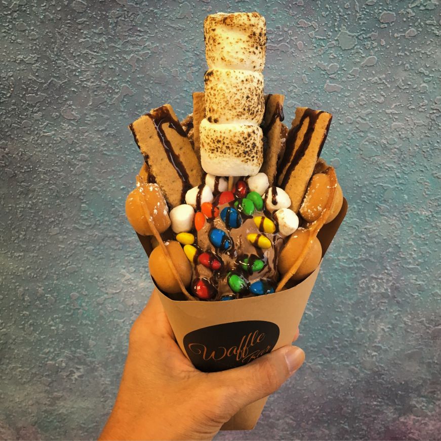 Hand holding rolled bubble waffle filled with chocolate ice cream and garnished with graham crackers, toasted marshmallows, and M&Ms