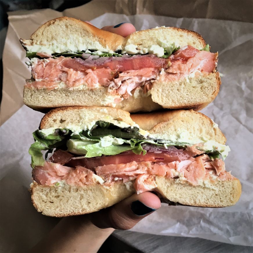 Hand holding a bagel sandwich piled with salmon, onions, tomatoes, and lettuce
