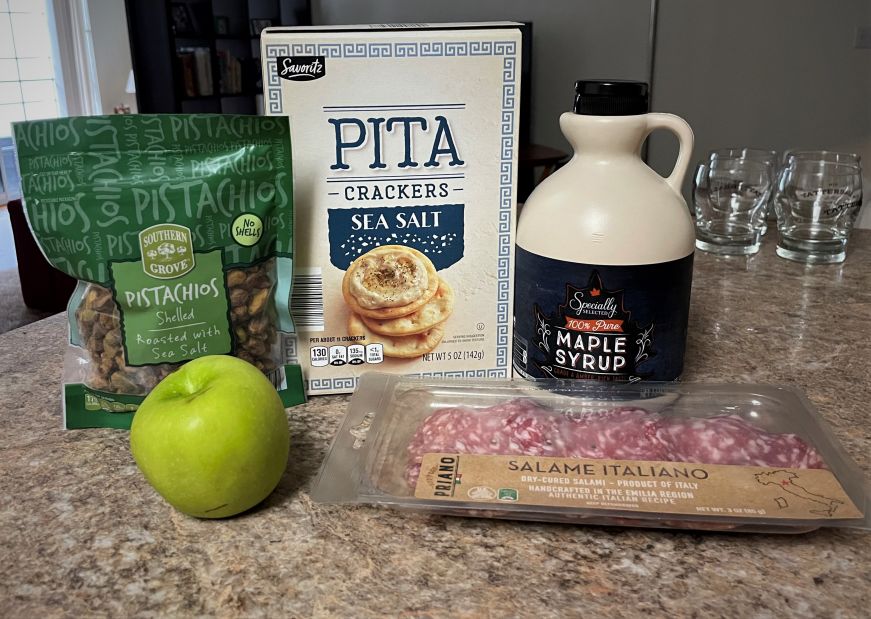 Granny smith apple, bag of pistachios, box of pita crackers, bottle of maple syrup, and package of salami arranged on kitchen counter