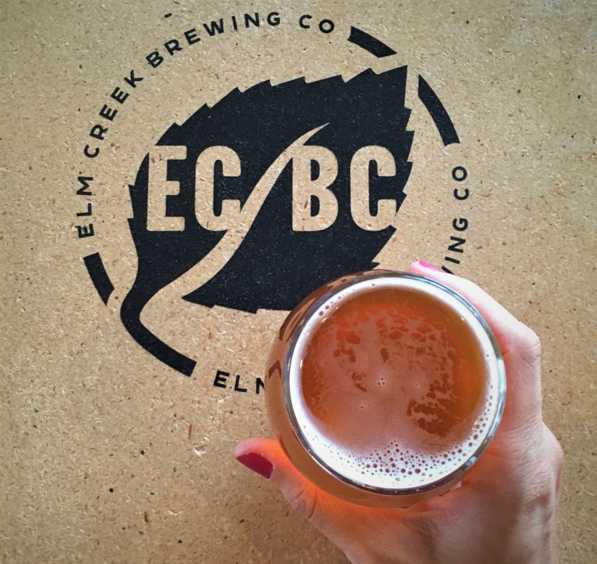 Top down view of hand holding glass of beer on a table painted with the logo for Elm Creek Brewing Co.