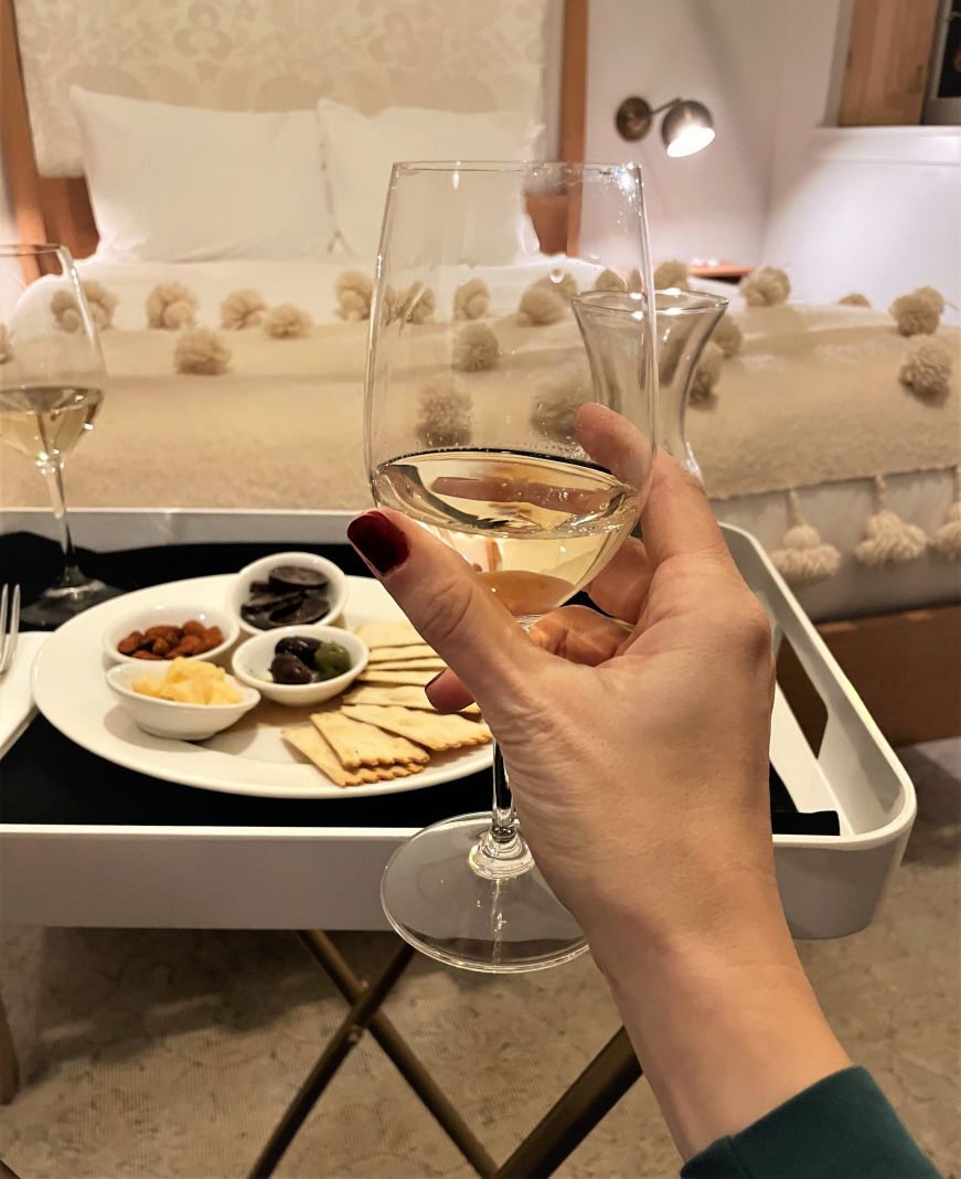 Hand holding glass of white wine with a tray of antipasti and bed in the background