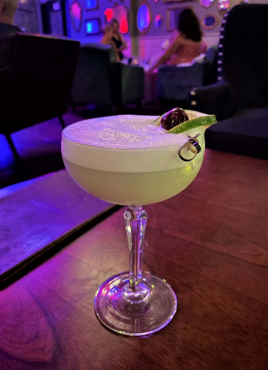 Coupe glass with a cocktail topped with egg white, a lime peel, and cherry