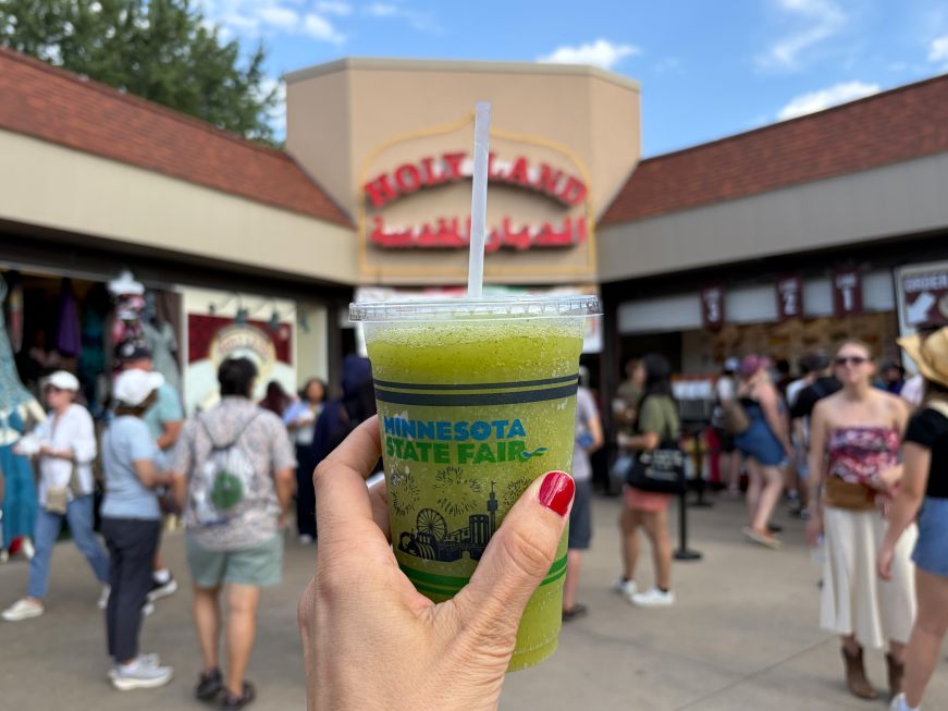 Hand holding a Mediterranean lemonade slushie in front of the sign for Holy Land