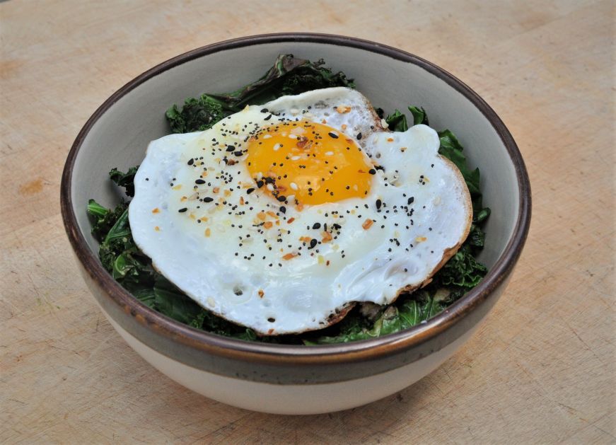 Bowl filled with sauteed kale and topped with a fried egg and everything bagel seasoning