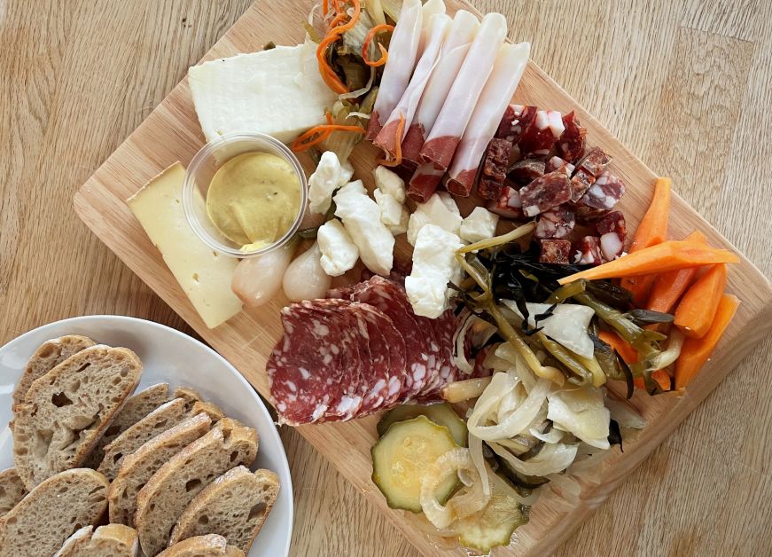 Serving board piled with cured meats, cheeses, and pickled vegetables, with a plate of sliced bread on the side