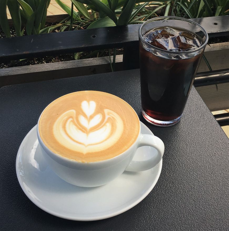 Latte with a flower pattern and a glass of cold brew coffee on an outdoor table with greenery in the background, Parallel, Minneapolis Latte with a flower pattern and a glass of cold brew coffee on an outdoor table with greenery in the background, Parallel, Minneapolis