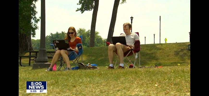 Screenshot of a news story showing Mike and Stacy sitting in lawn chairs in a park and working on laptops