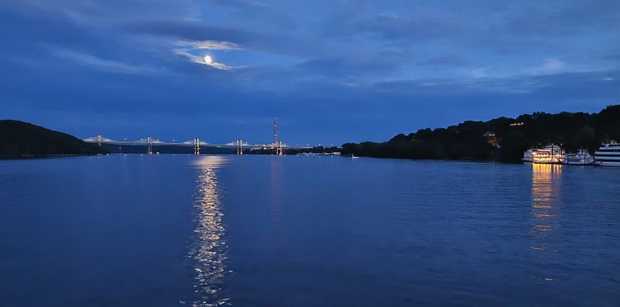 Nighttime view of a bridge in the distance with a full moon and its reflection