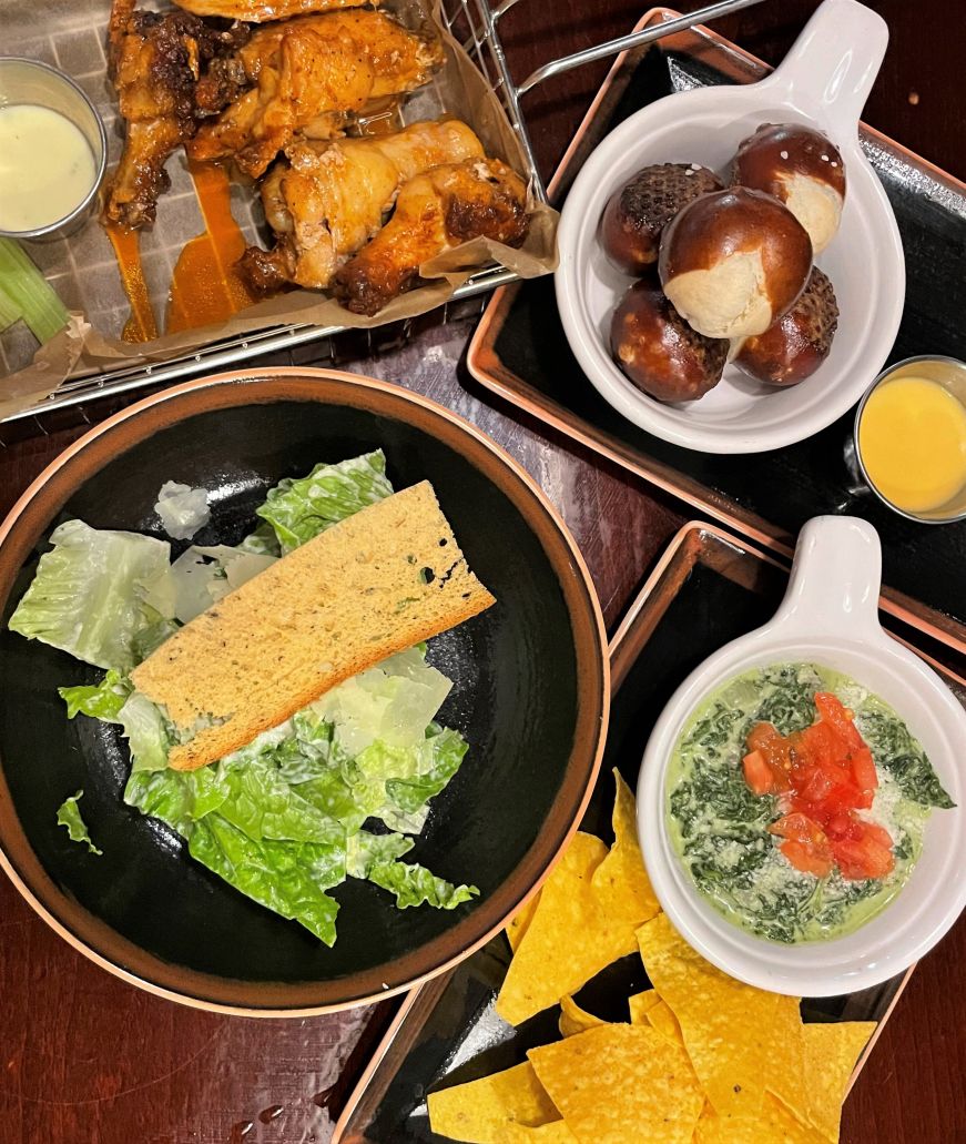 Top down view of plates of wings, caesar salad, pretzel bites, and spinach dip with tortilla chips