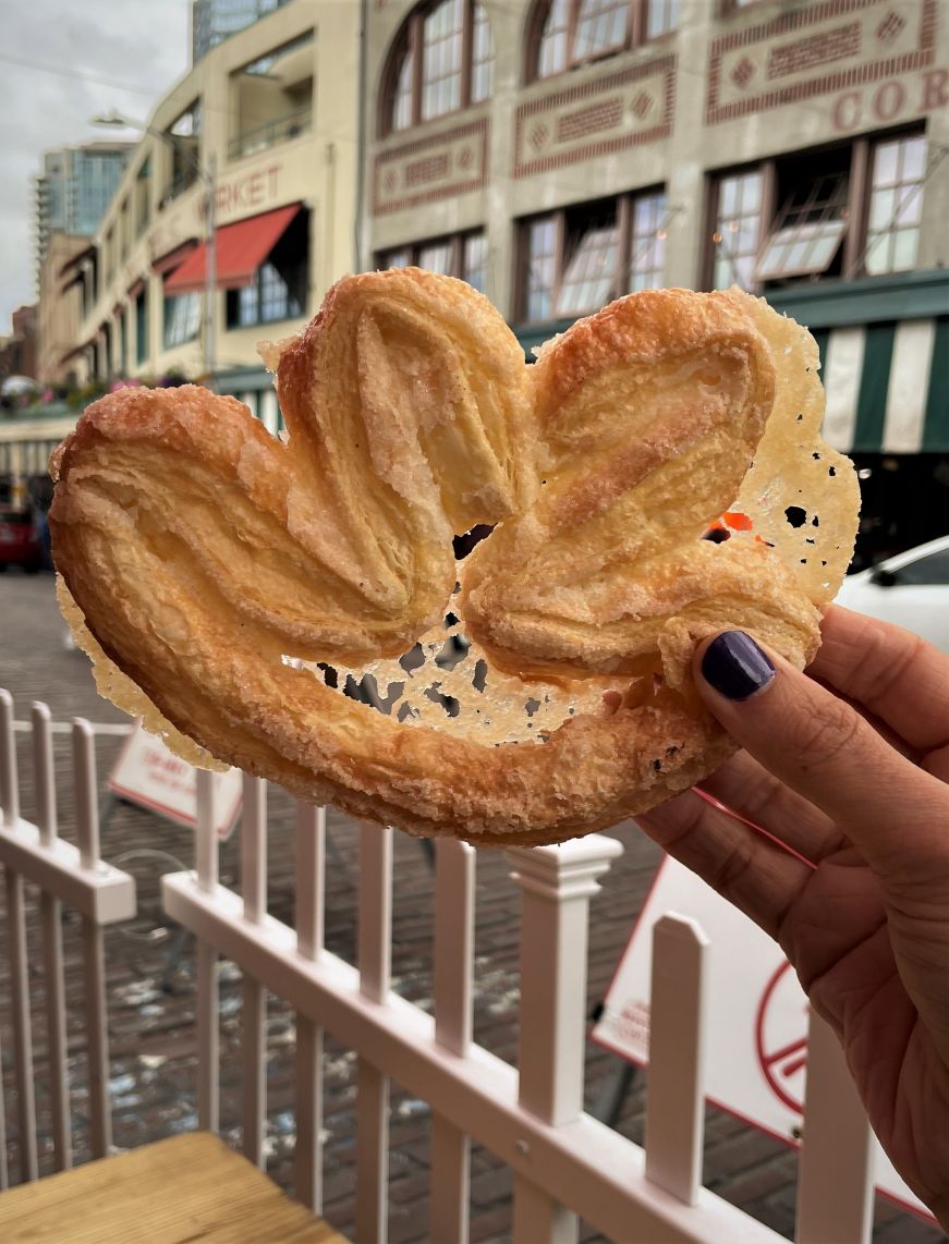 Hand holding a palm-shaped pastry with historic Pike Place Market buildings in the background