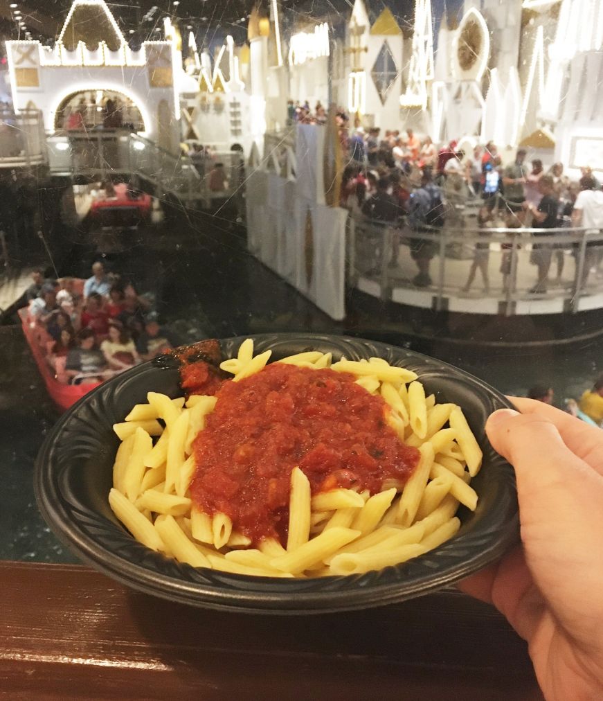 Hand holding a bowl of penne pasta topped with marinara sauce with Disney's It's A Small World Ride in the background