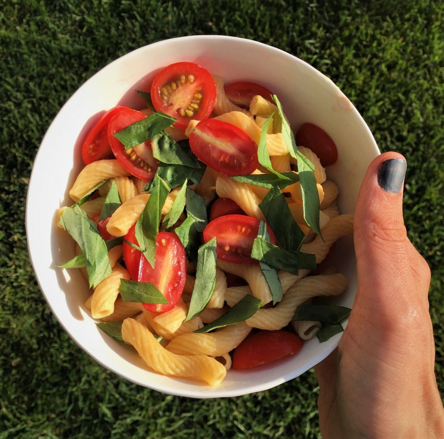 Hand holding white bowl with spiral pasta, chunks of fresh tomato, and chopped fresh basil