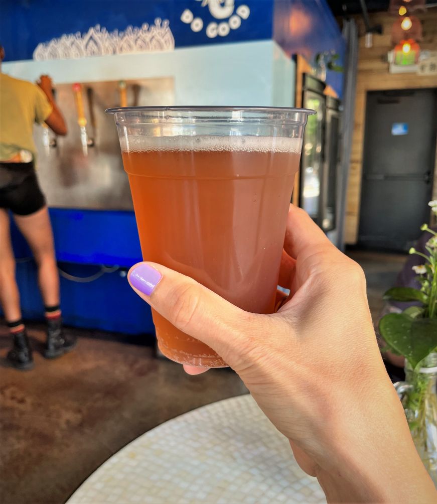 Stacy's hand holding a pint glass filled with orange colored kombucha