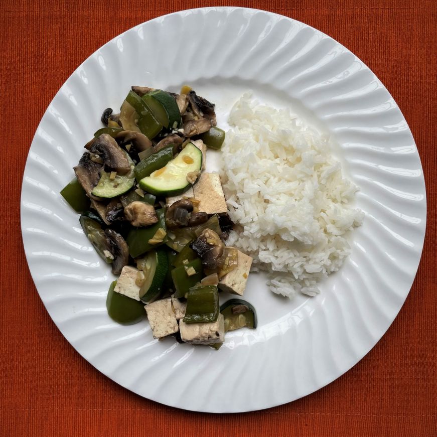 White plate with a portion of white rice and a stir fry with green peppers, zucchini, mushrooms, and tofu